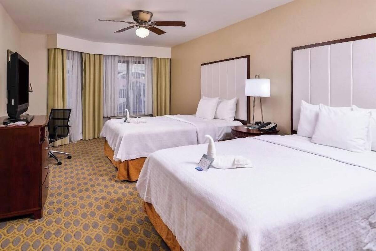 Une chambre d’hôtel avec deux lits, un bureau, une télévision, un ventilateur de plafond et une fenêtre avec des rideaux.