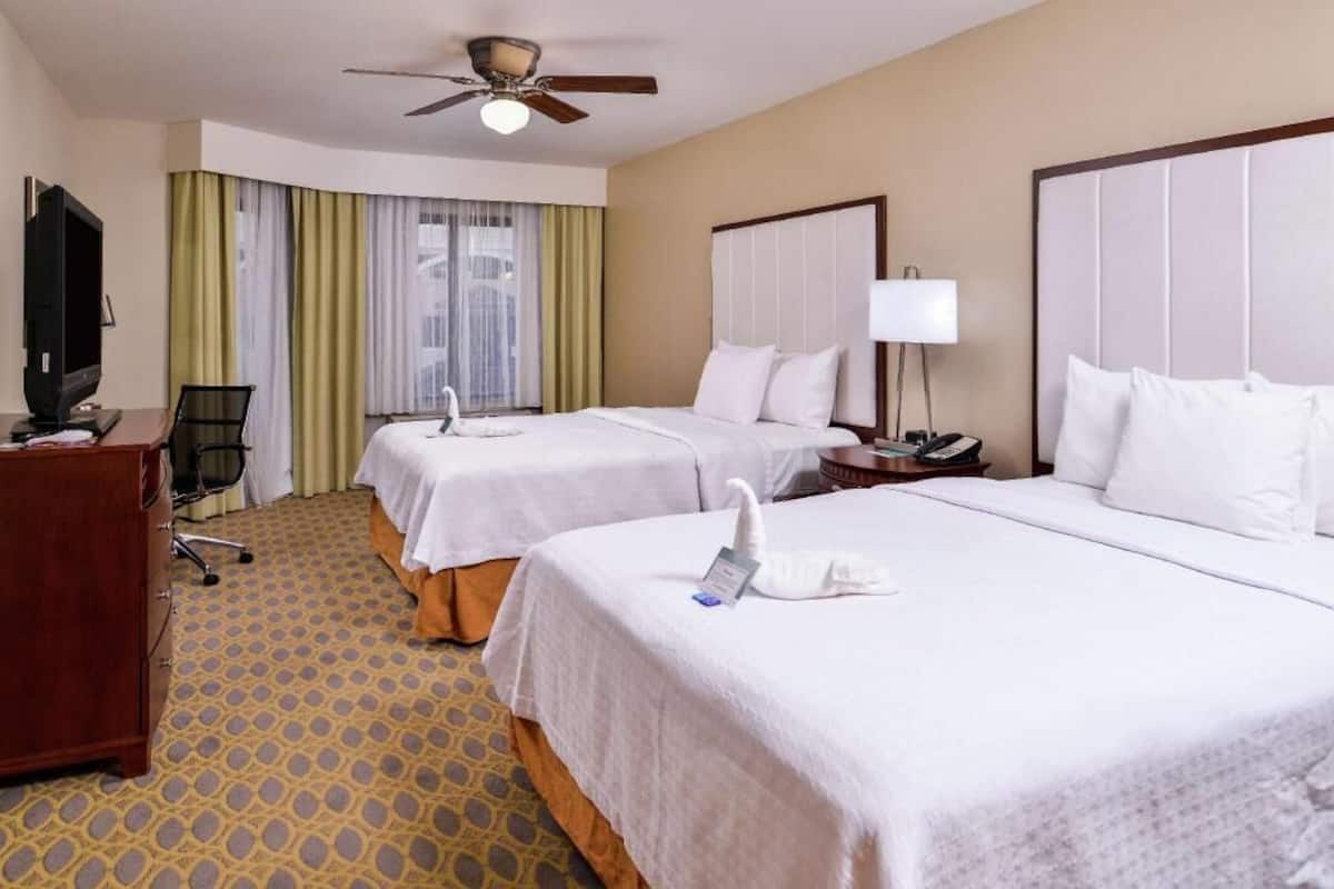 Une chambre d’hôtel avec deux lits, un bureau avec une télévision, un ventilateur de plafond et une fenêtre avec des rideaux.