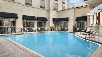 A swimming pool with lounge chairs and umbrellas, surrounded by a building with large windows and a tiled floor.