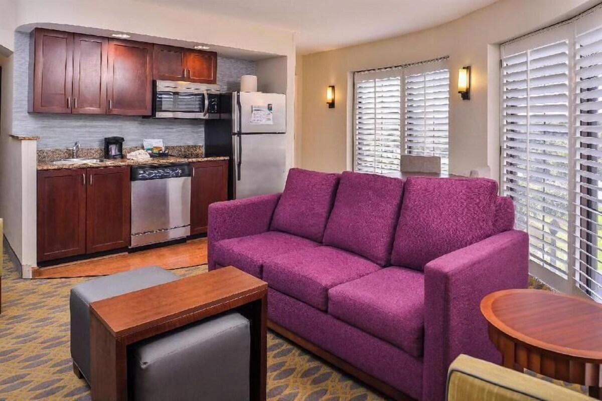 A compact living space with a kitchenette, a purple sofa, a small wooden table, and a patterned carpet.
