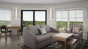 A modern living room with a grey sofa, a wooden coffee table, and a small table with chairs. There are large windows with blinds, and a balcony with a view. - Homewood Suites by Hilton Jacksonville Downtown-Southbank (Jacksonville)