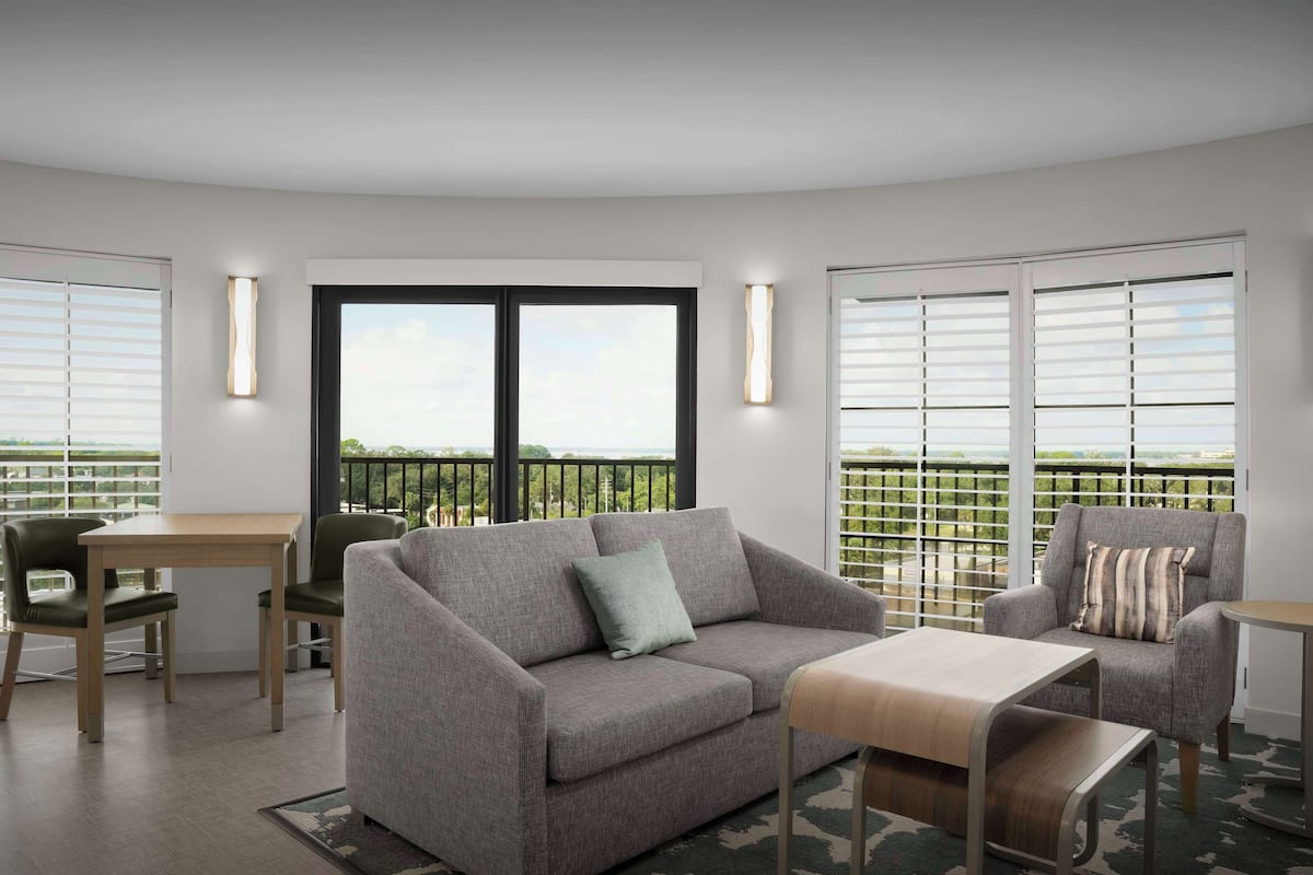 Un salon moderne avec un canapé gris, une table basse en bois et une petite table accompagnée de chaises. De grandes fenêtres sont équipées de stores, et un balcon offre une vue dégagée.