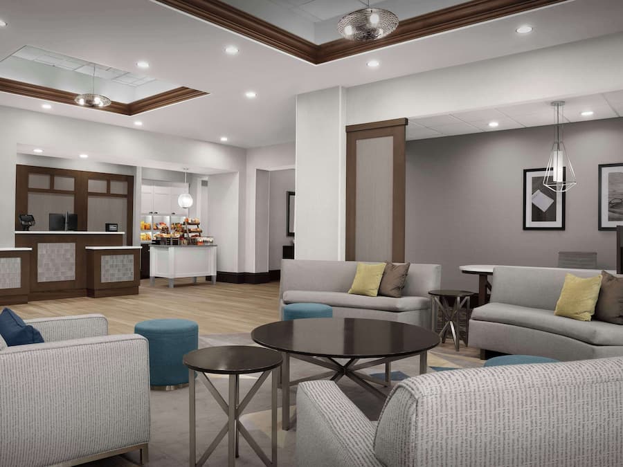 A modern hotel lobby with a reception desk, seating area, and a kitchen in the background.