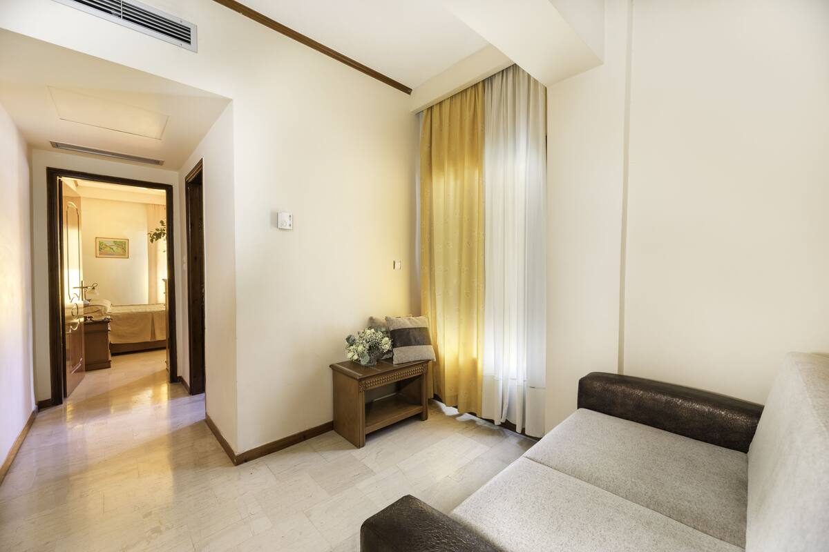 A living room with a sofa, a small side table with a lamp, and a view of a bedroom through an open door.