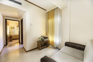 A living room with a sofa, a small side table with a lamp, and a view of a bedroom through an open door. - Famissi Hotel (Kalabaka)