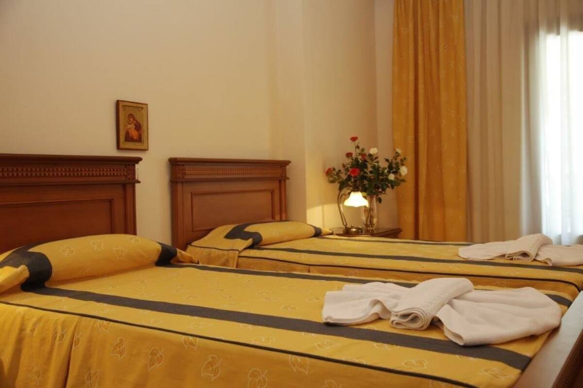 A hotel room with two beds, each with yellow bedding and black trim. There is a bedside table with a lamp and a vase of flowers.