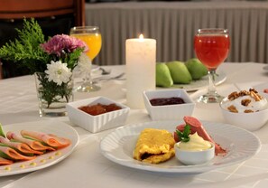 A table set with various dishes, including a plate with sliced ham and vegetables, a plate with a rolled omelette, and a glass of red drink. There are also small bowls with condiments and a candle on the table. - Famissi Hotel (Kalabaka)