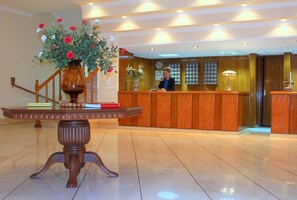 A hotel reception area with a wooden reception desk, a large vase with flowers, and a polished floor. - Famissi Hotel (Kalabaka)