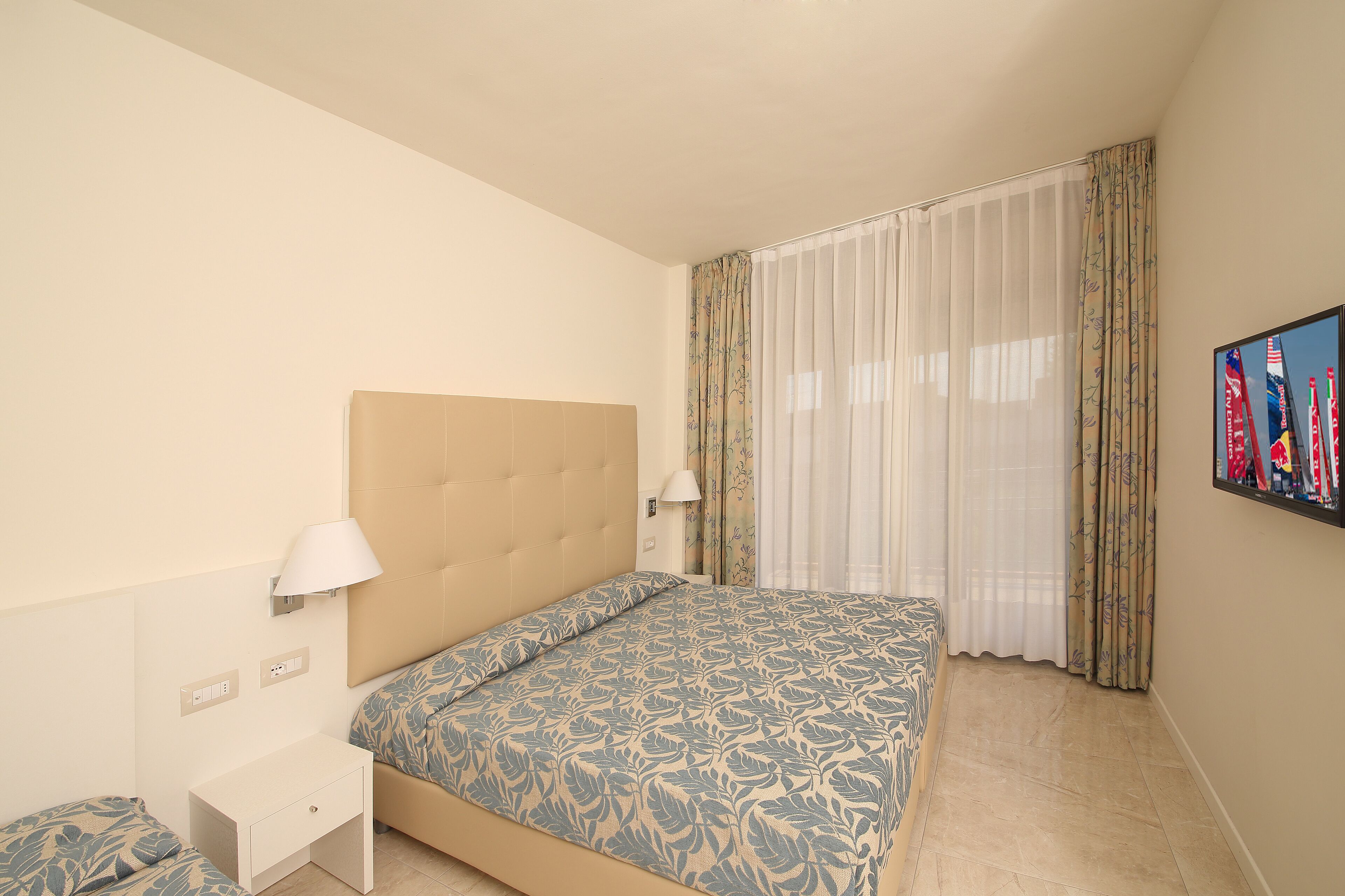Una habitación de hotel con una cama, una mesita de noche, una lámpara y una ventana con cortinas.