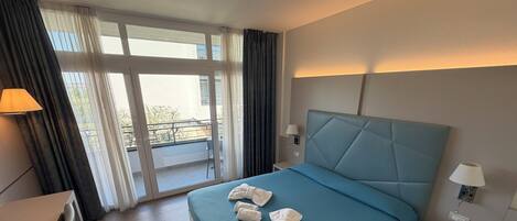 Habitación de hotel con cama, escritorio, silla, mesita de noche con teléfono y vista al exterior a través de un ventanal grande.