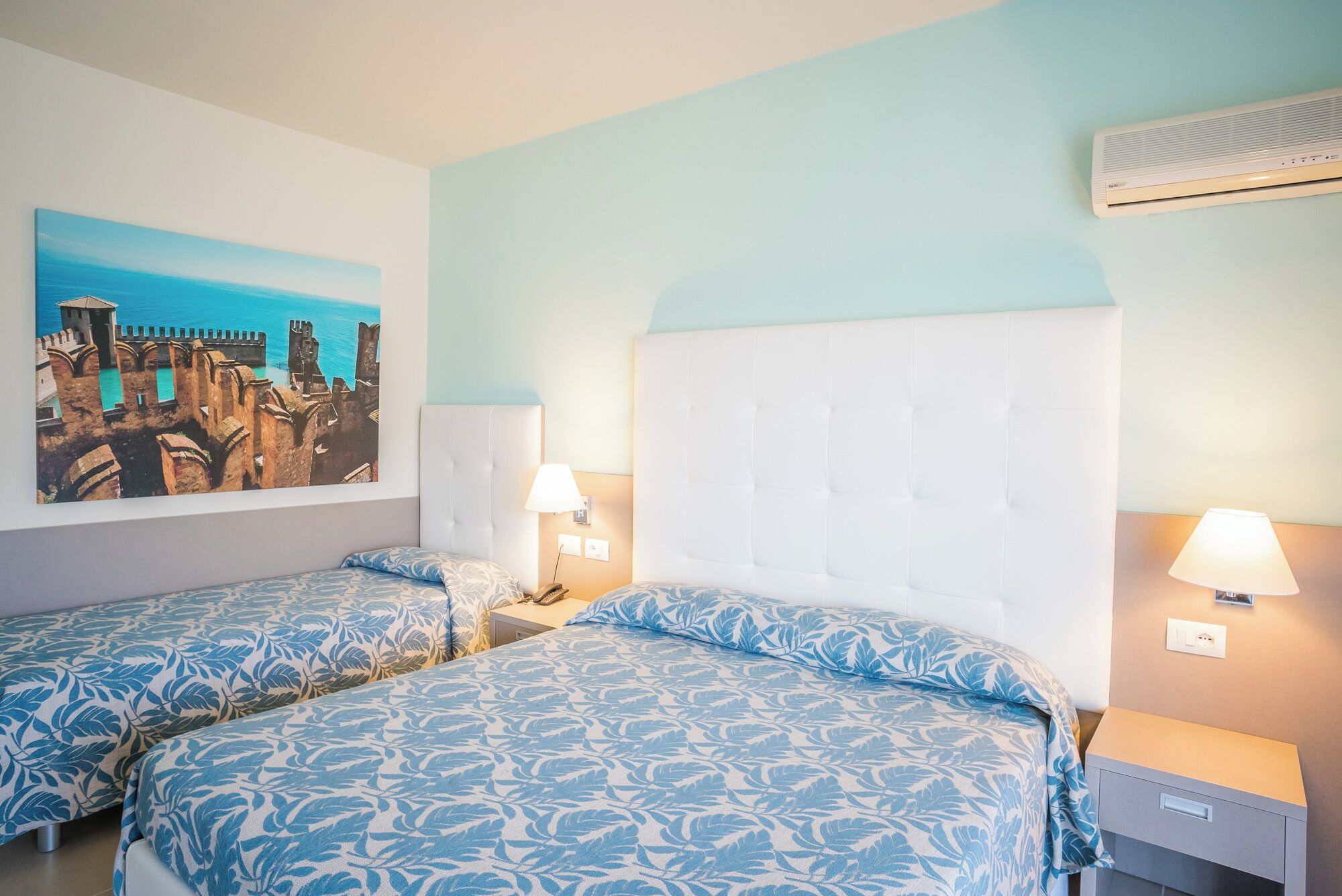 A hotel room with two beds, a large painting of a coastal scene, and an air conditioning unit.