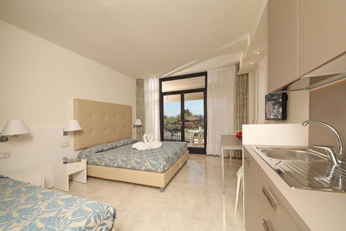 A hotel room with a bed, bedside table, lamp, and a view of the outdoors.