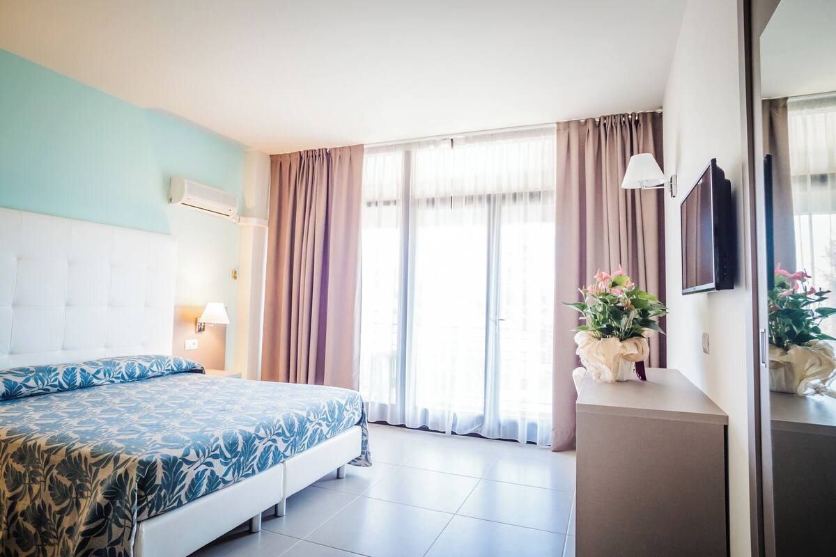 A hotel room with a bed, a bedside table with a lamp, a TV mounted on the wall, and a window with curtains.
