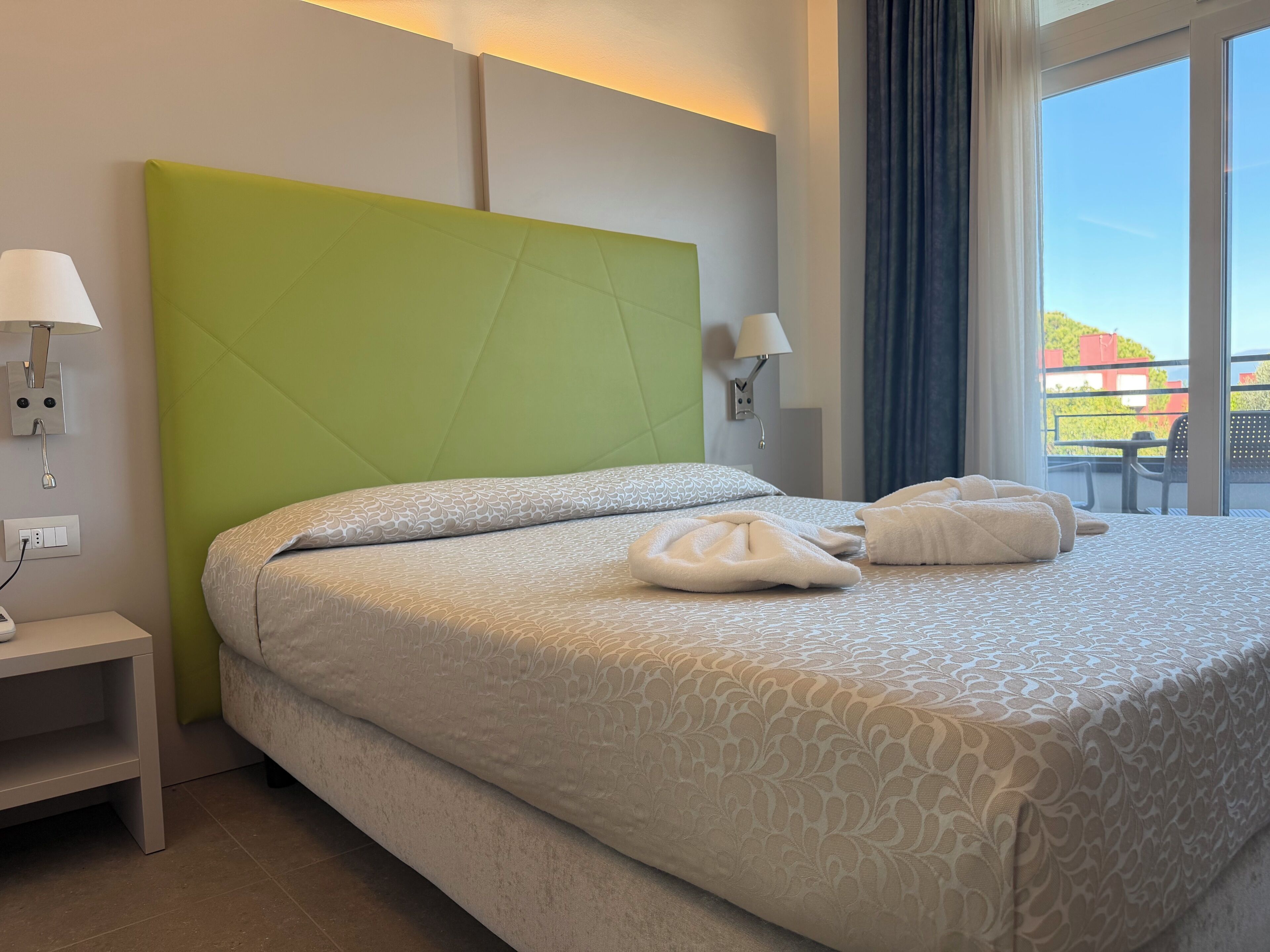 A hotel room with a green headboard, a bed with patterned bedding, two bedside lamps, and a view of the outdoors.