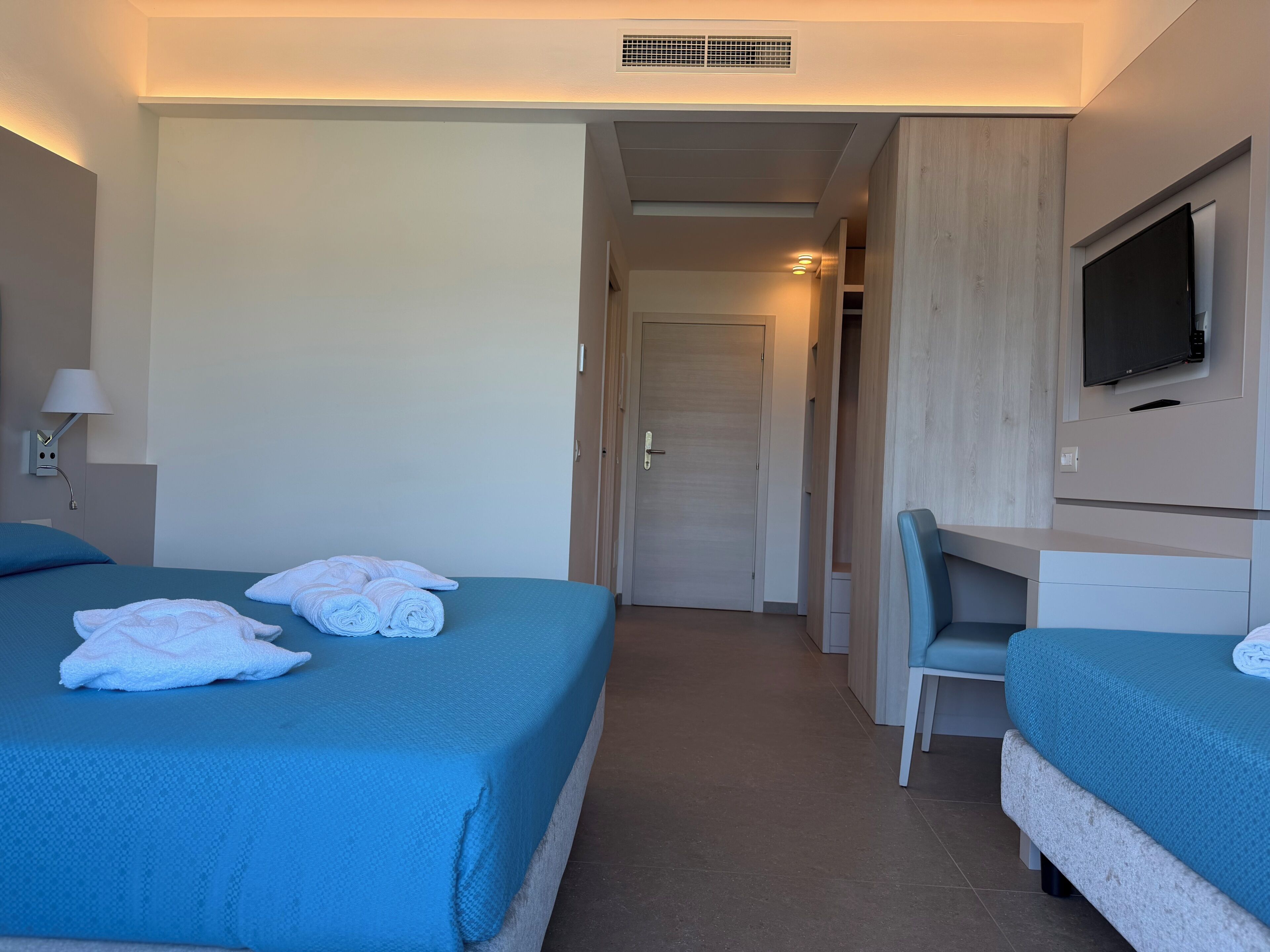 A hotel room with two beds, a desk, a chair, a TV, and a door leading to a hallway.