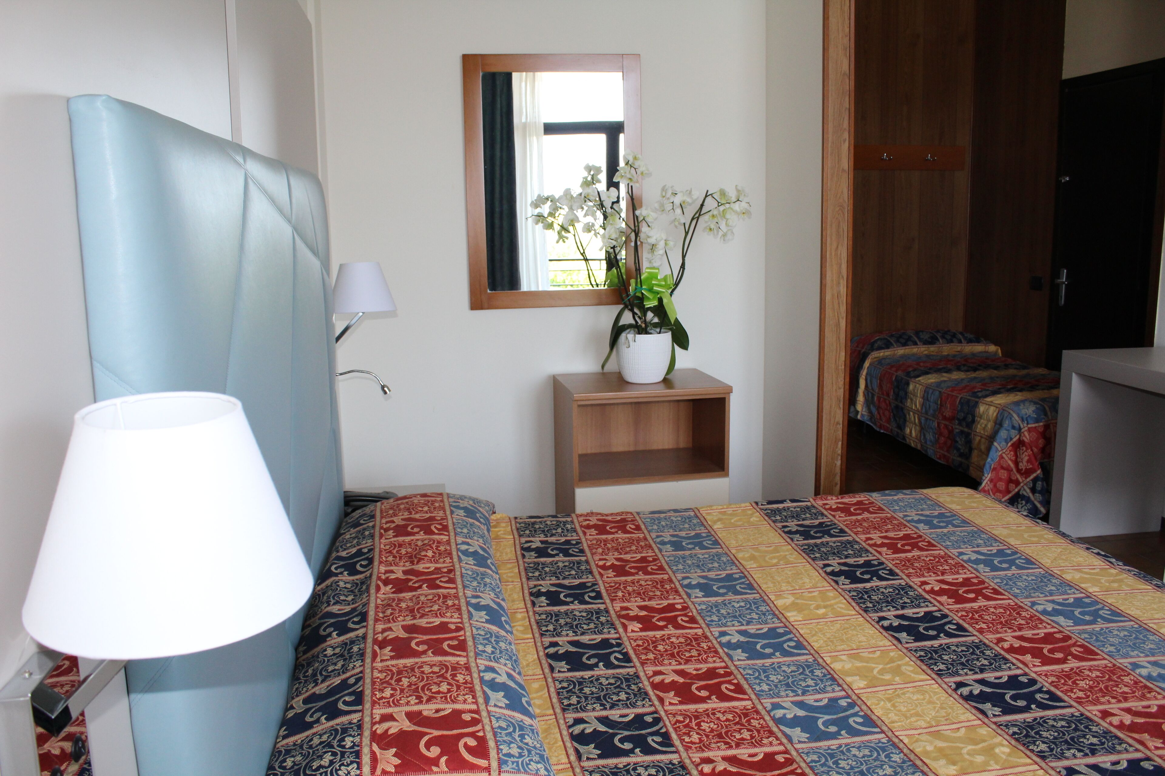 Une chambre à coucher avec un grand lit, une table de chevet avec une lampe, une petite table de nuit en bois, une plante en pot et un miroir au mur.