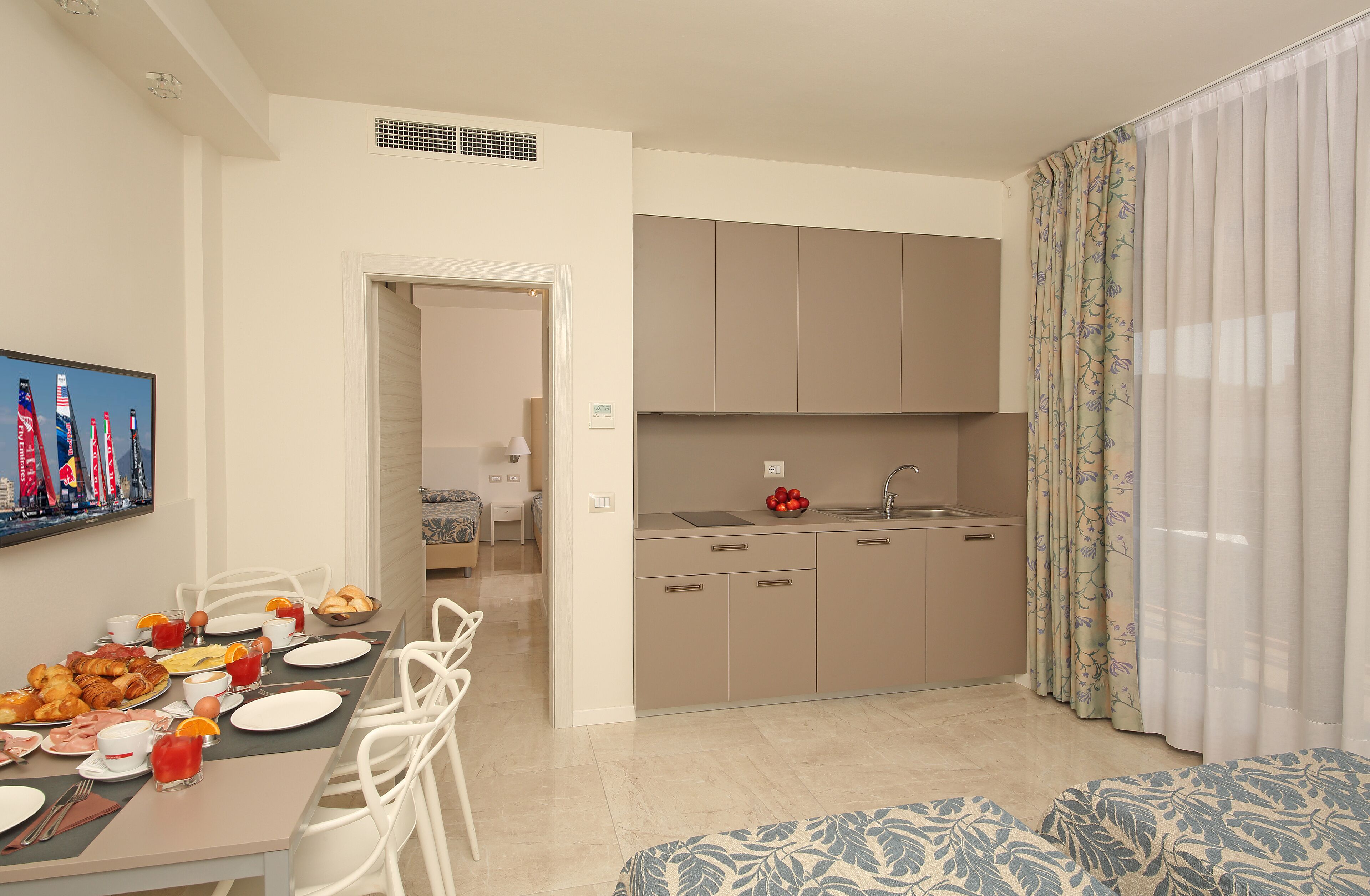 A modern kitchen with a dining area, a sofa, and a TV displaying a cityscape.