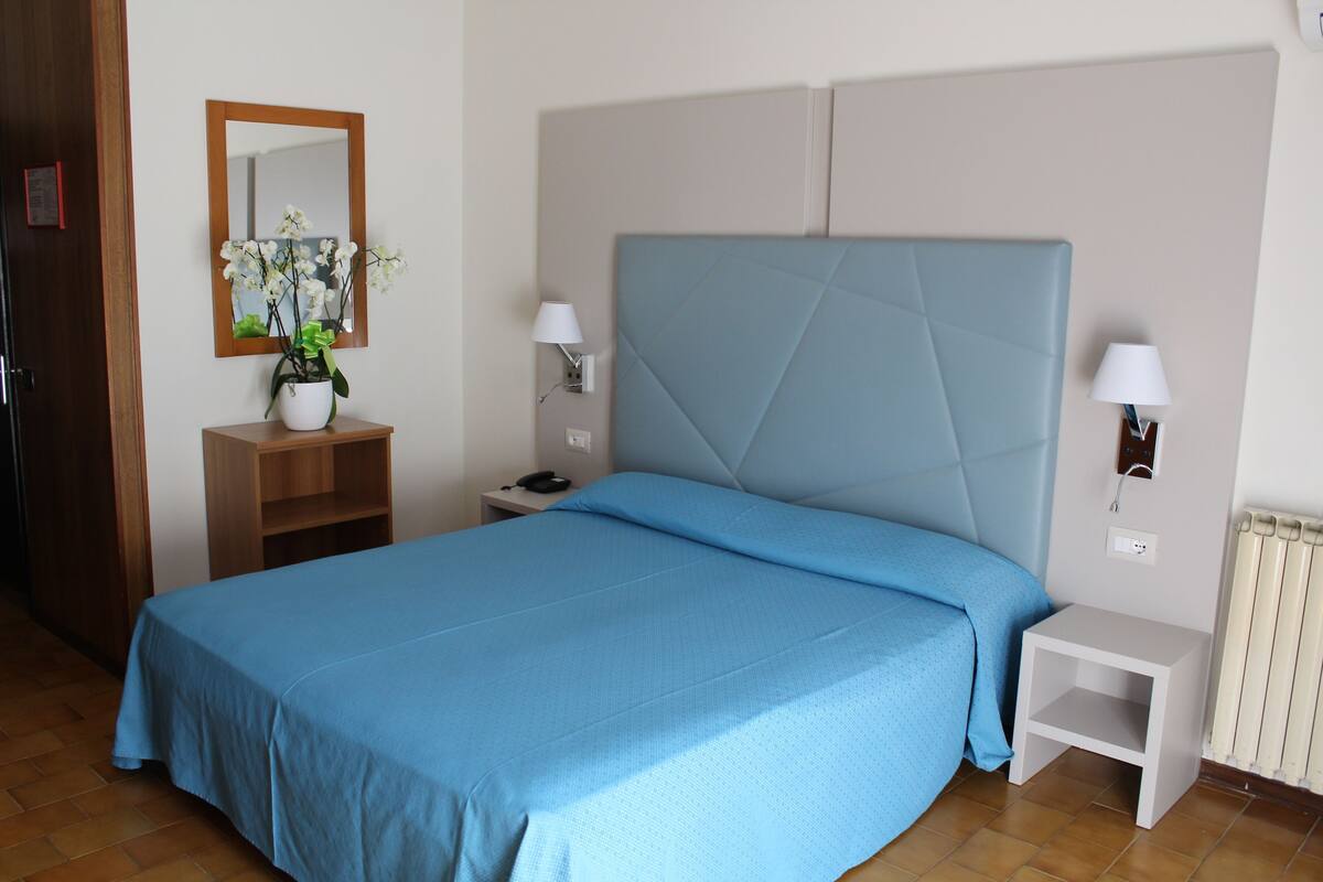 A bedroom with a bed, a nightstand, a lamp, a mirror, and a potted plant.