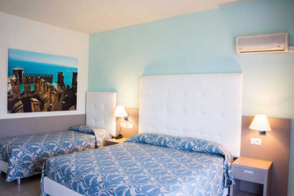 A hotel room with two beds, a large painting of a coastal cityscape, and an air conditioning unit on the wall.