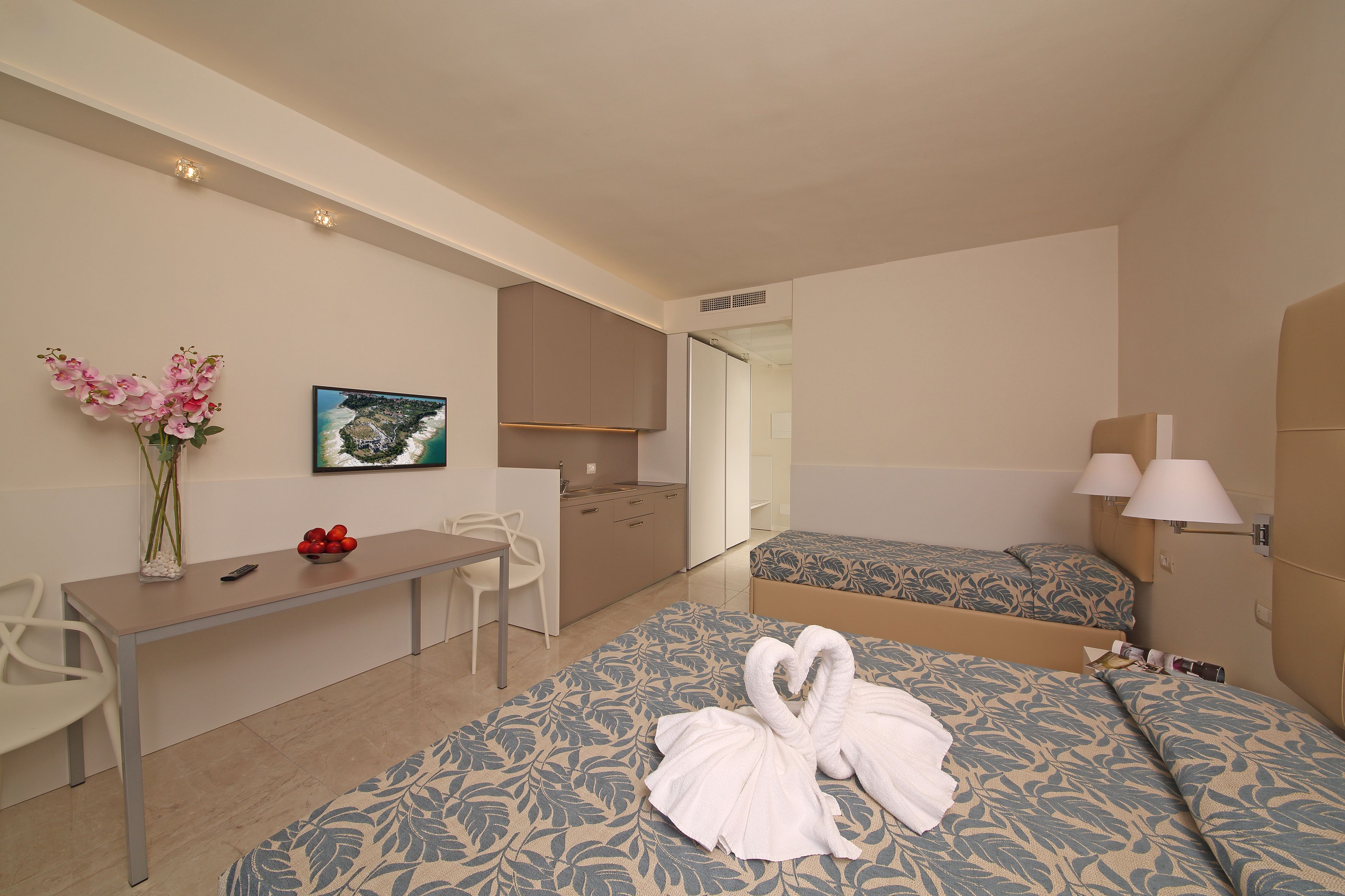 A hotel room with two beds, a desk, a chair, a small table with a vase of flowers, and a picture on the wall.