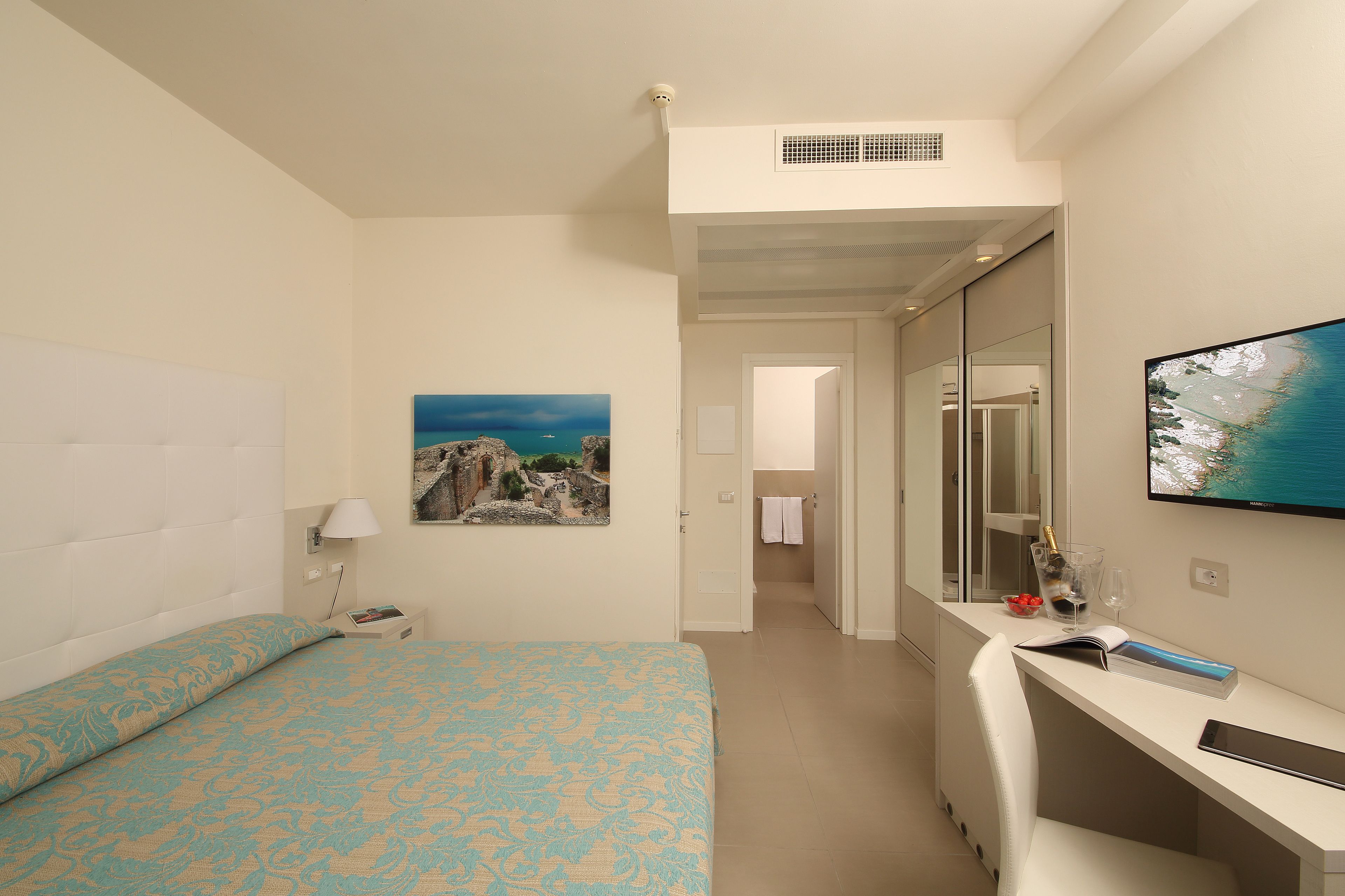 A hotel room with a bed, a desk, a television, and a painting on the wall.