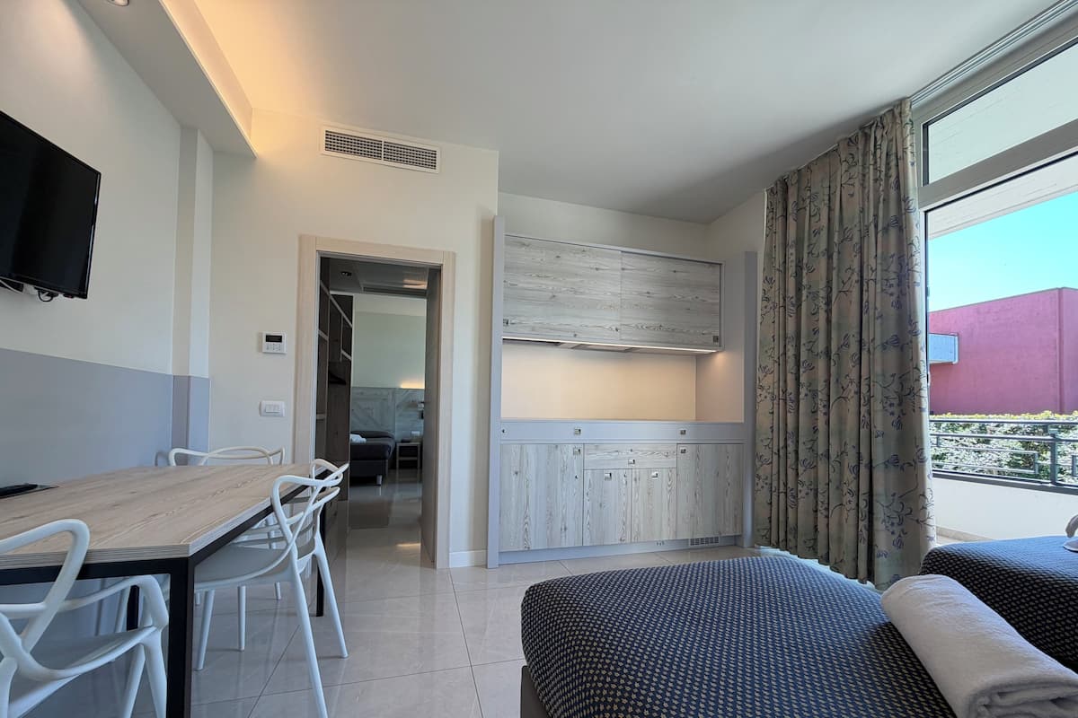 A hotel room with a bed, a desk with chairs, a kitchenette, and a view of the outside.