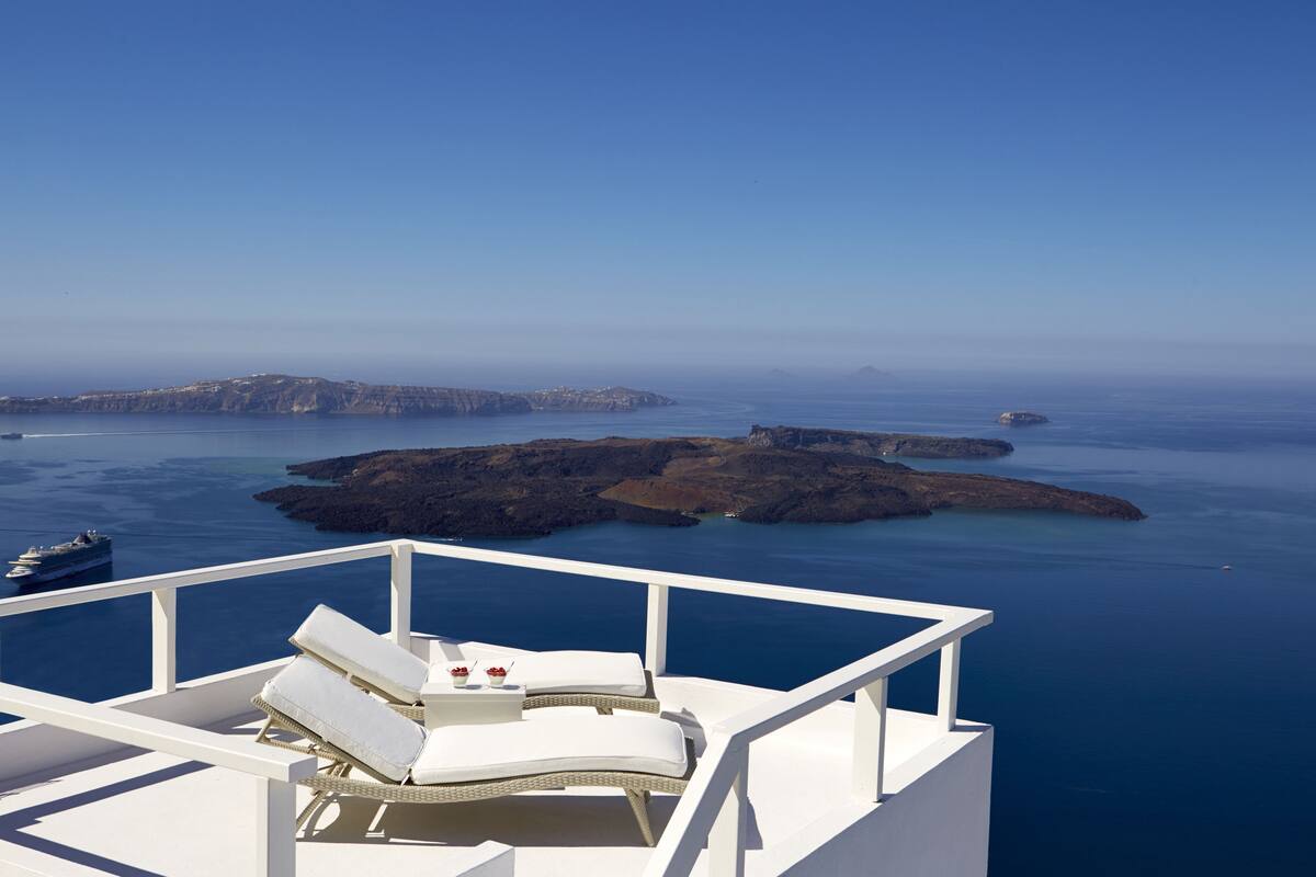 A balcony with white furniture overlooking a coastal landscape with rocky islands and a clear blue sea.