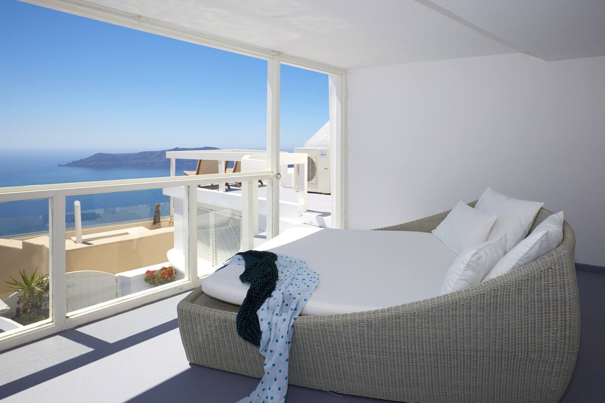 A bedroom with a large bed, white walls, and a balcony with a view of the sea.
