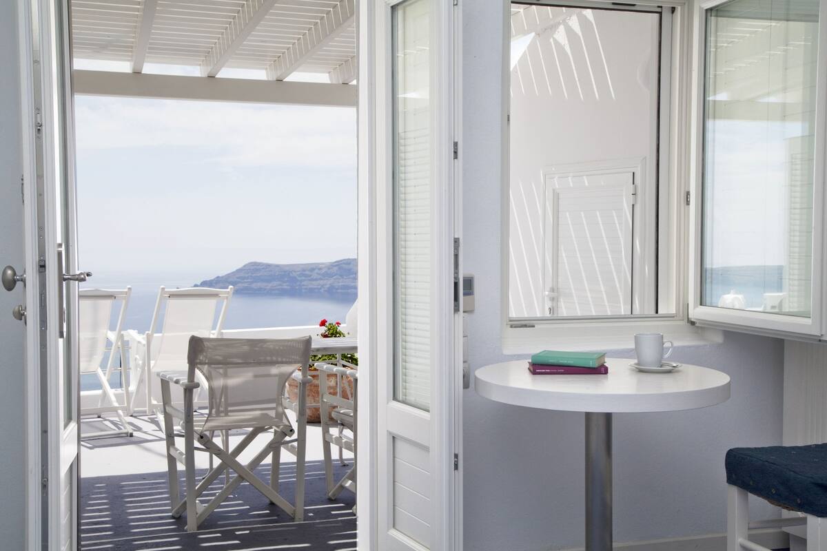 An open door leading to a balcony with white chairs, a small table with a cup and books, and a view of the sea and distant land.