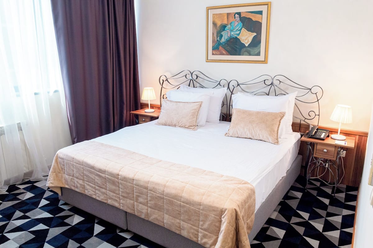 A neatly made bed with a beige bedspread and pillows, a metal headboard, bedside lamps, and a framed painting on the wall.