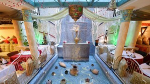 Un décor intérieur luxueux avec une fontaine centrale, des colonnes richement décorées et un sol orné de motifs.