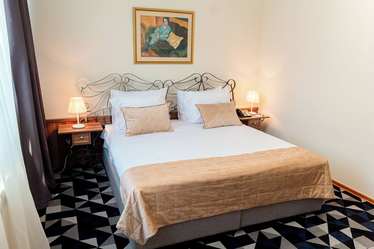 A hotel room with a bed, two bedside tables with lamps, a painting on the wall, and a patterned floor.