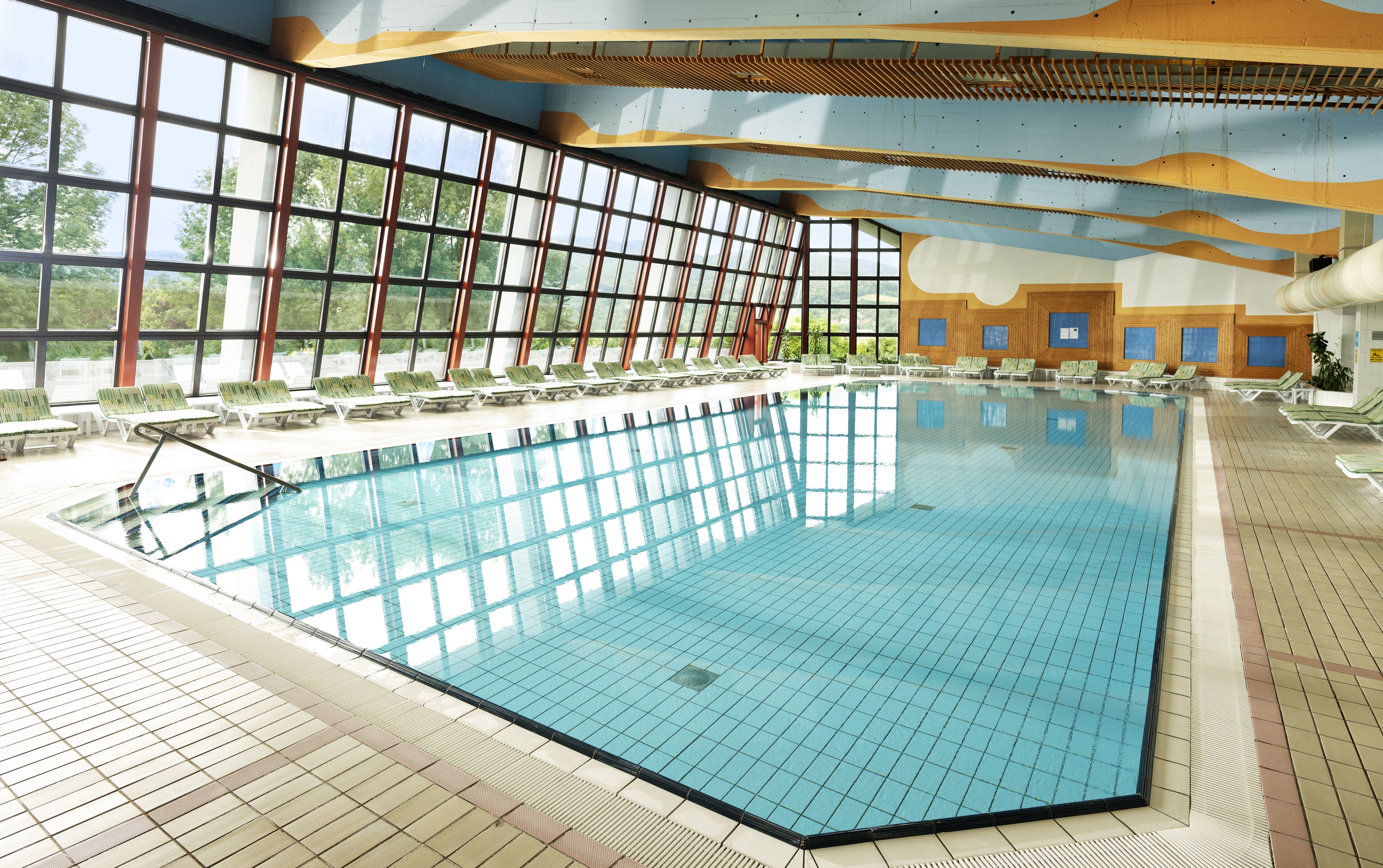 Une piscine intérieure à l’eau d’un bleu cristallin, entourée de chaises longues et dotée de grandes fenêtres qui offrent une vue sur la verdure environnante.