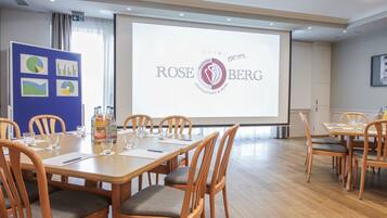 A conference room with a large screen displaying the Rose Berg hotel logo, wooden tables, and chairs.