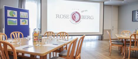 A conference room with a large screen displaying the Rose Berg hotel logo, wooden tables, and chairs.