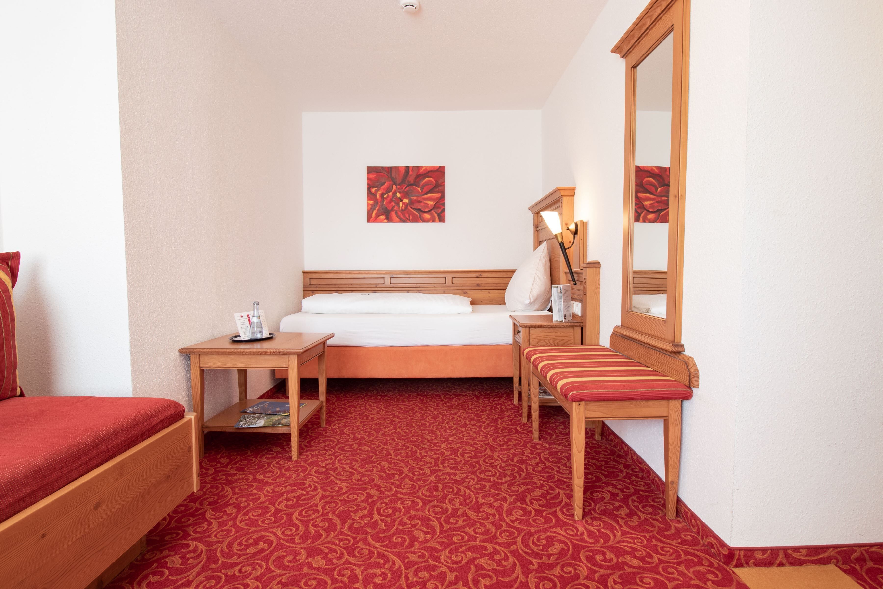 Hotelkamer met een bed, nachtkastje met een lamp, een bankje, een spiegel en een rood karpet met een patroon.