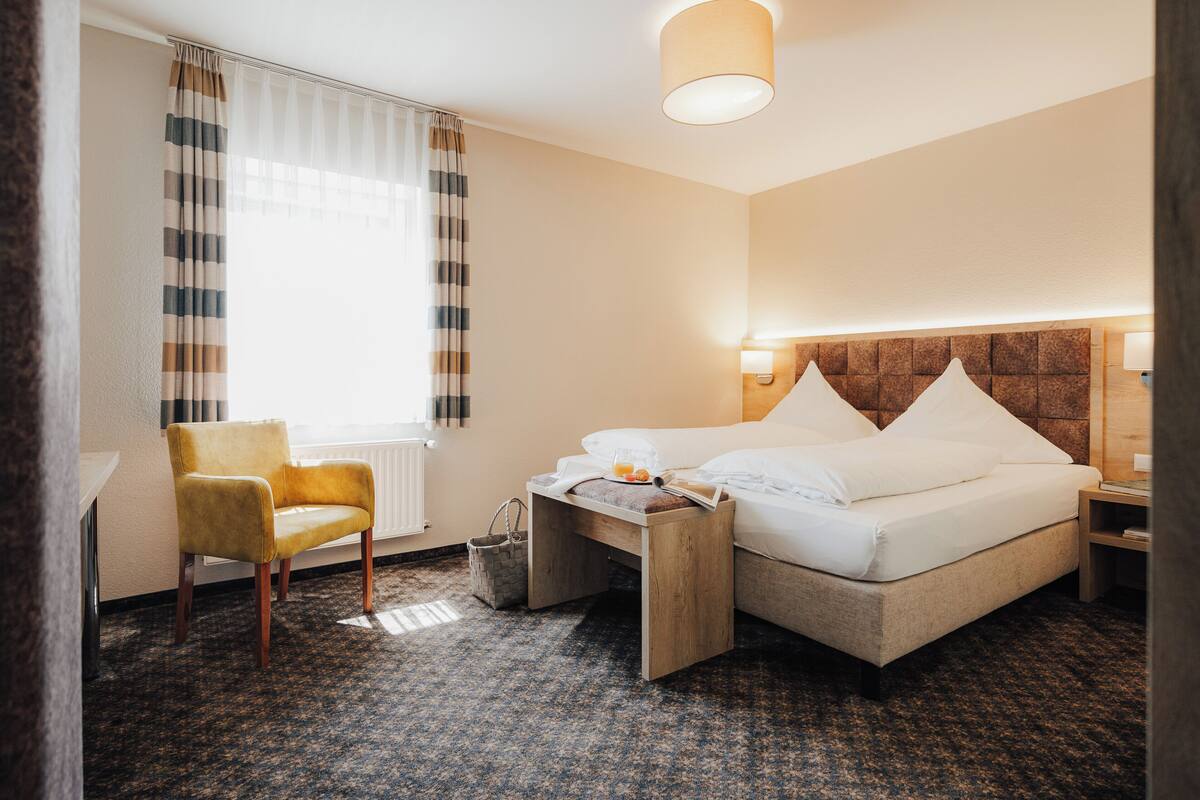 Ein Hotelzimmer mit einem Bett, einem Sessel, einem Nachttisch und einem Fenster mit Vorhängen.