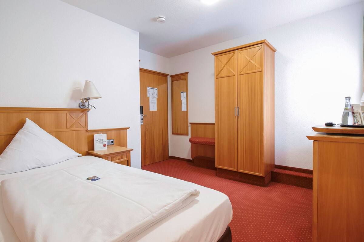 Ein Hotelzimmer mit Bett, Holzschrank, Schreibtisch mit Lampe und rotem Teppichboden.