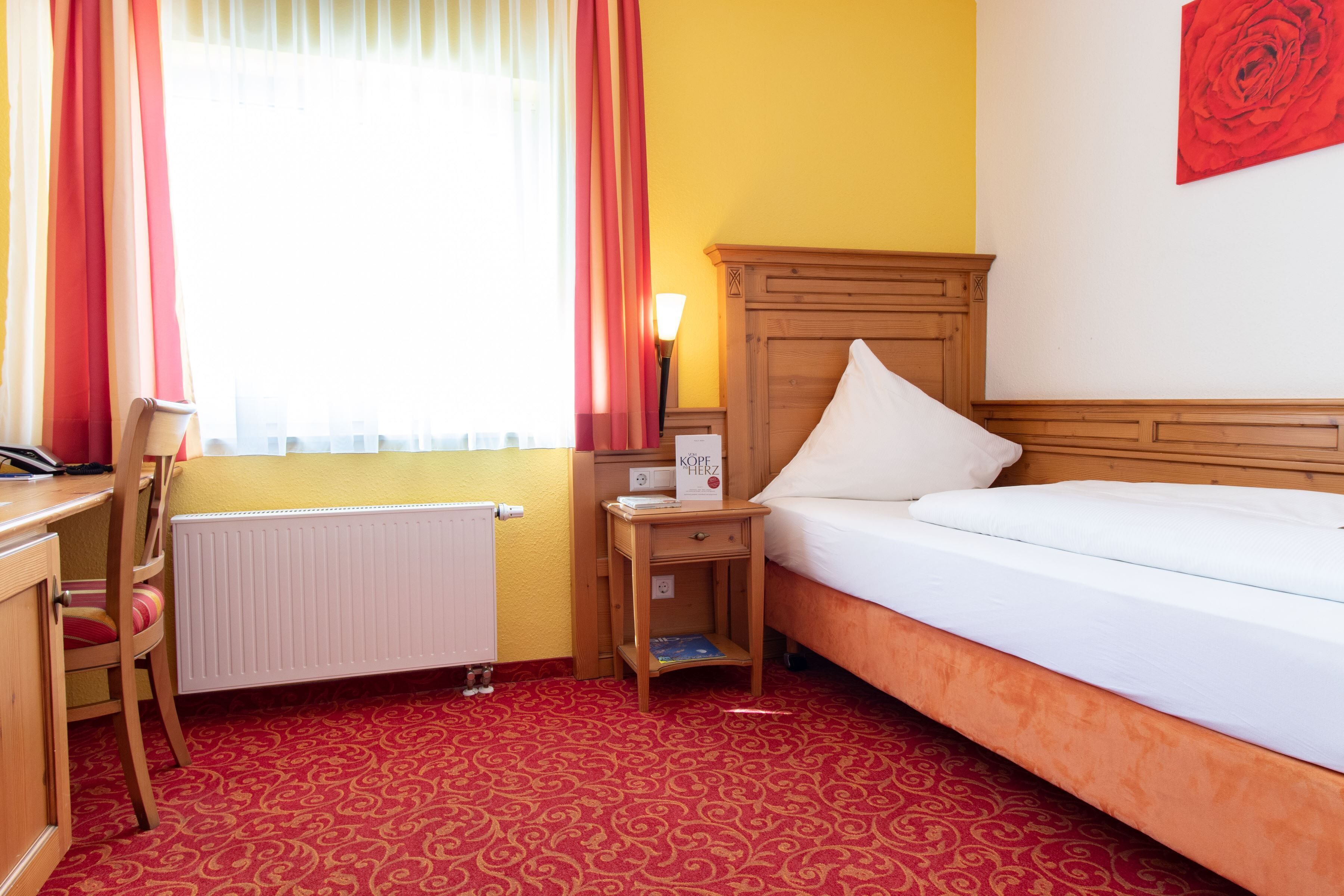 A hotel room with a wooden bed, a desk, a chair, a nightstand with a lamp, and a radiator.