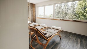 Ein Raum mit Holzbettplattformen für Massagen, einem großen Fenster mit Blick auf Bäume und einem Holzboden.
