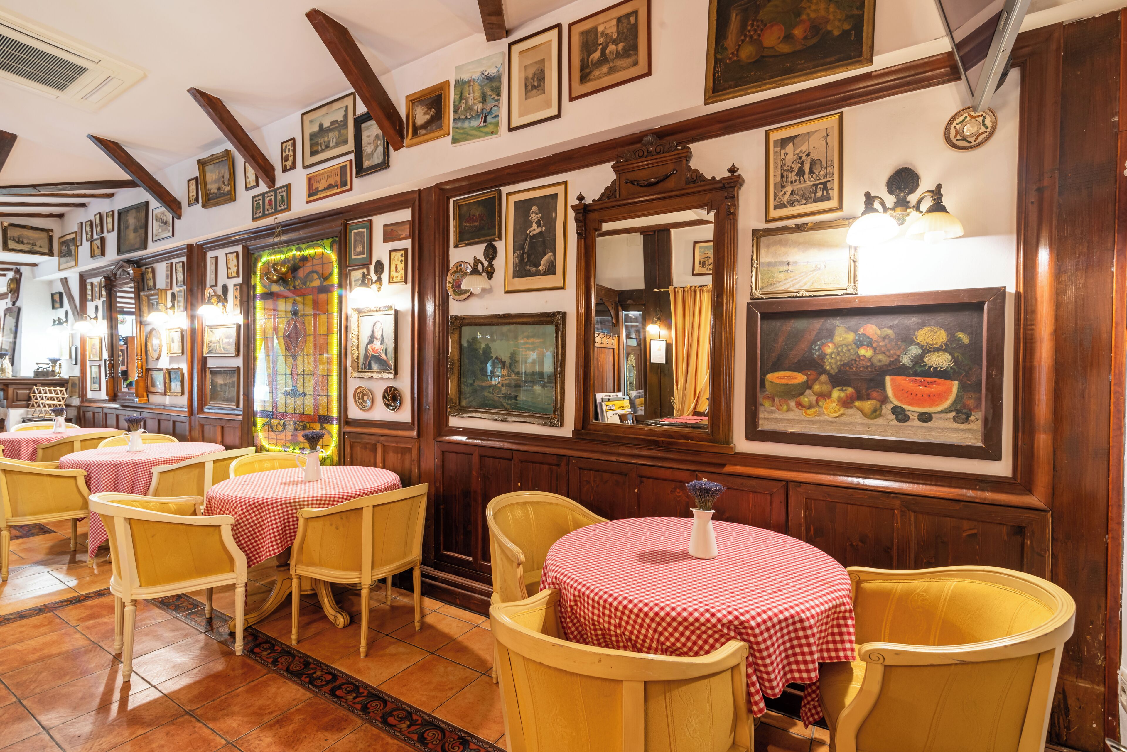 A restaurant interior with wooden beams, red and white checkered tablecloths, and various framed artworks on the walls.