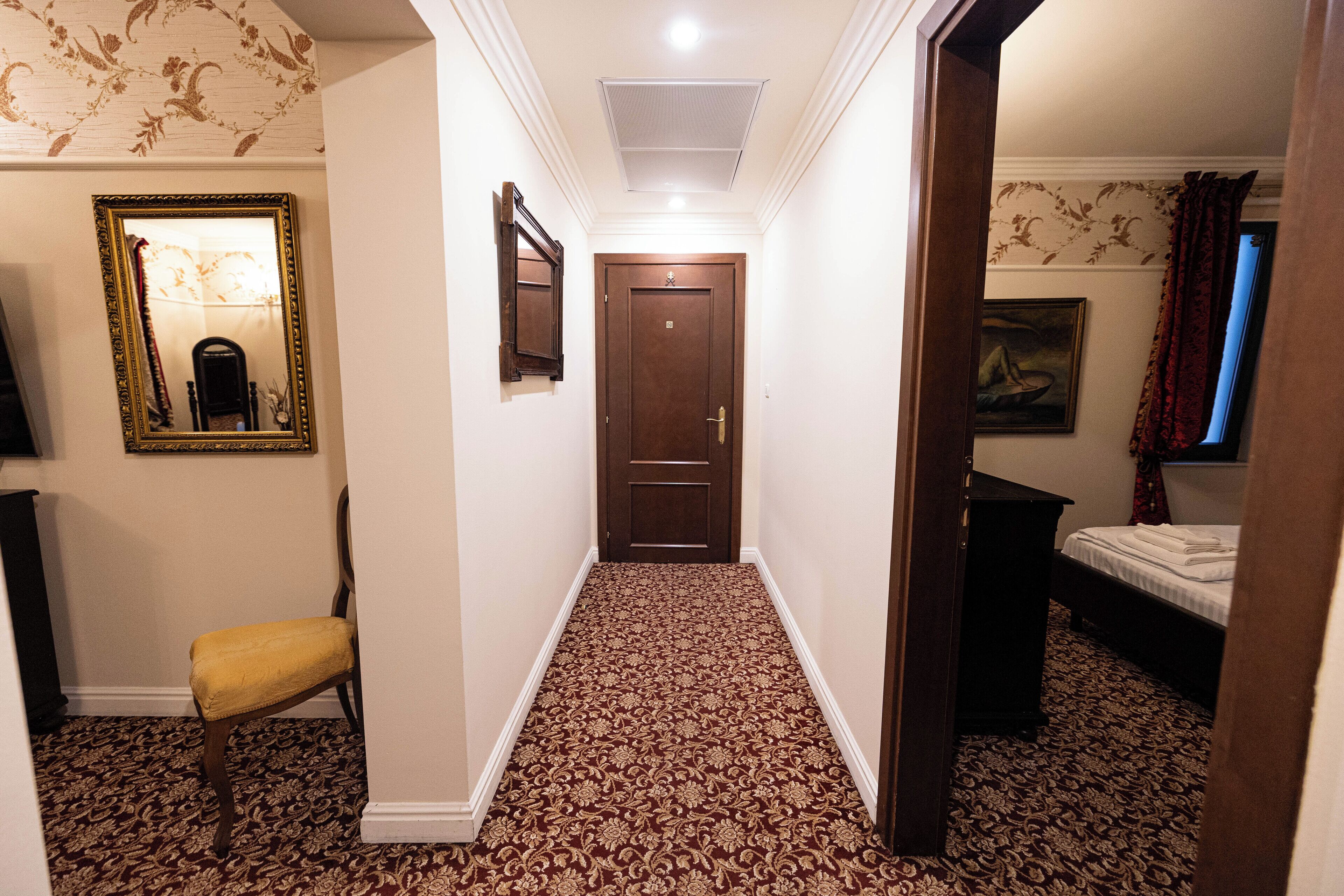 Un pasillo estrecho con alfombra estampada, una puerta de madera, una silla y una cama en la habitación contigua.