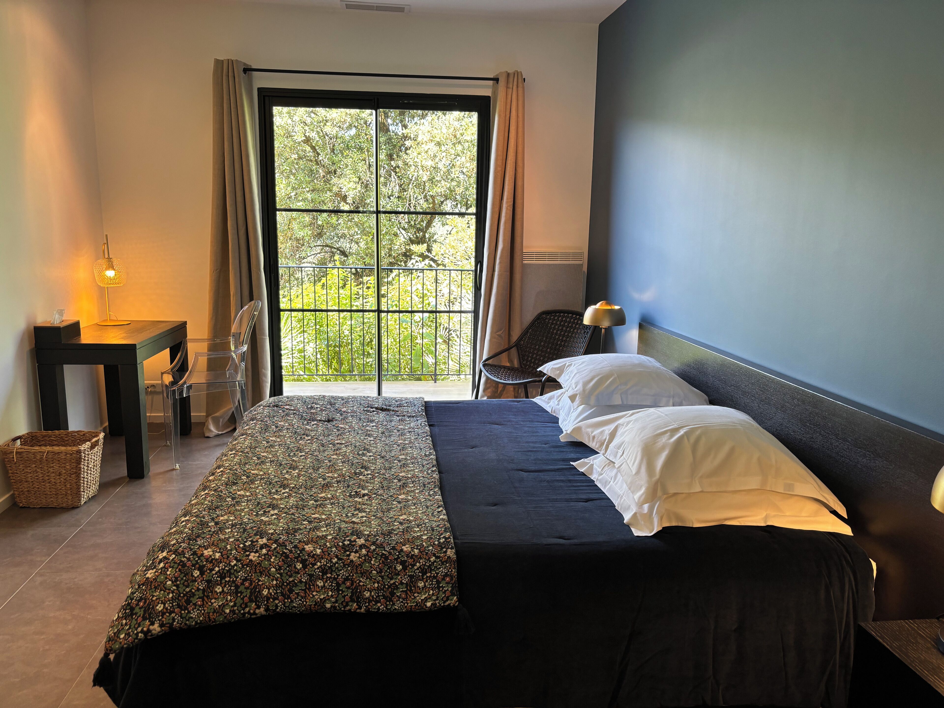 Ein Schlafzimmer mit einem Bett, einem Sessel, einem Tisch und Blick ins Grüne durch ein Fenster.