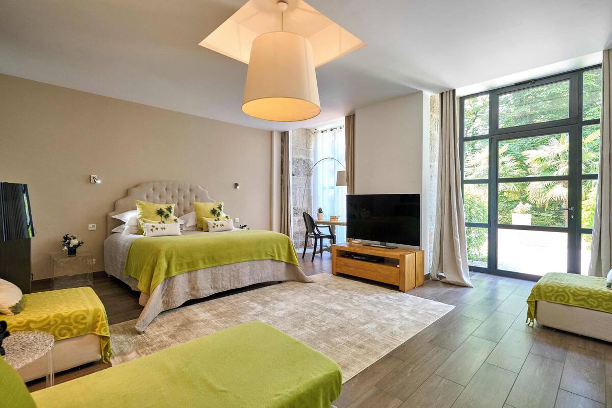 A bedroom with a large bed, a television, and a window with a view of greenery.