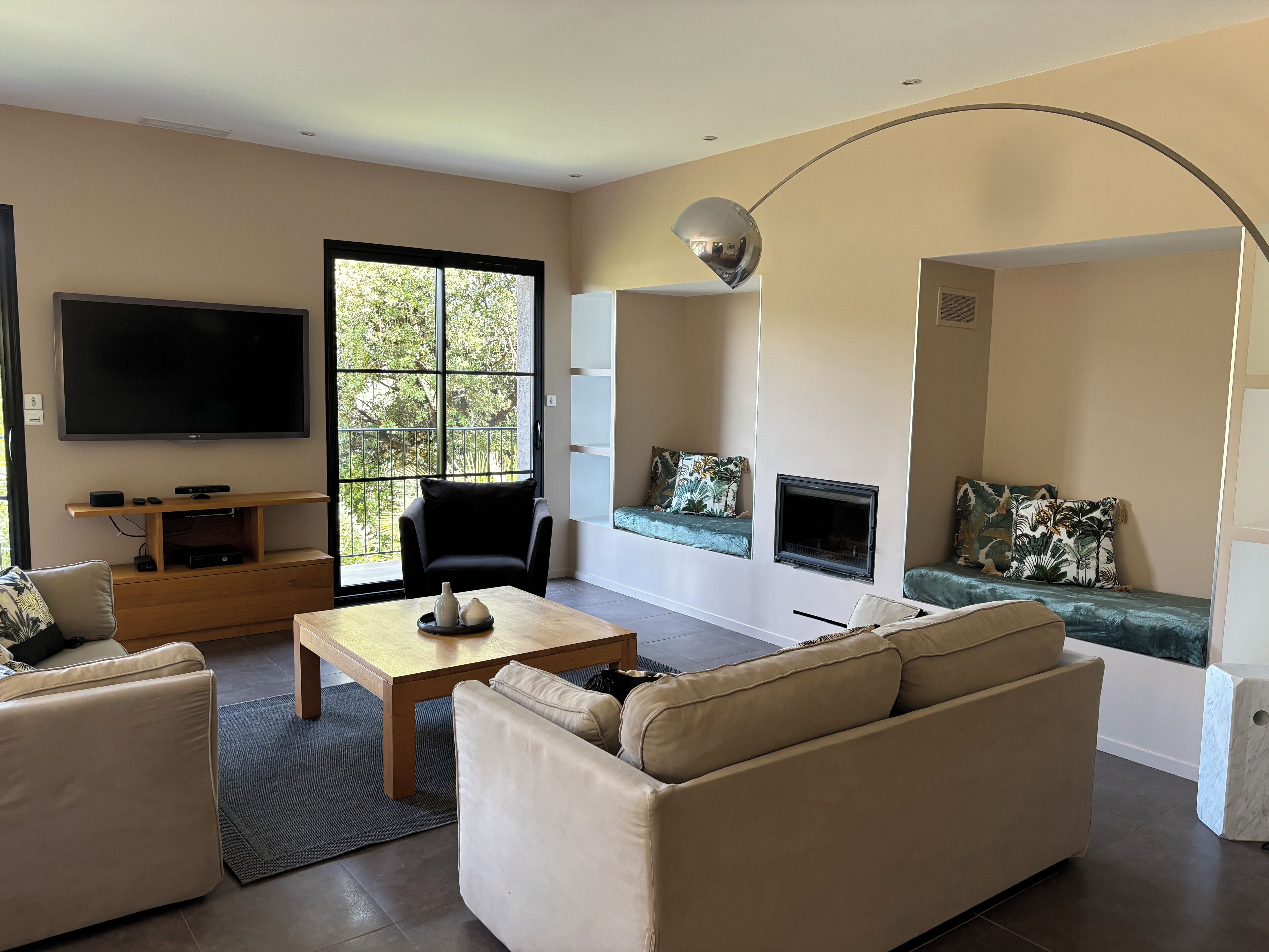 A modern living room with a flat-screen TV, a wooden coffee table, beige sofas, a black armchair, a fireplace, and a large window with a view of trees.