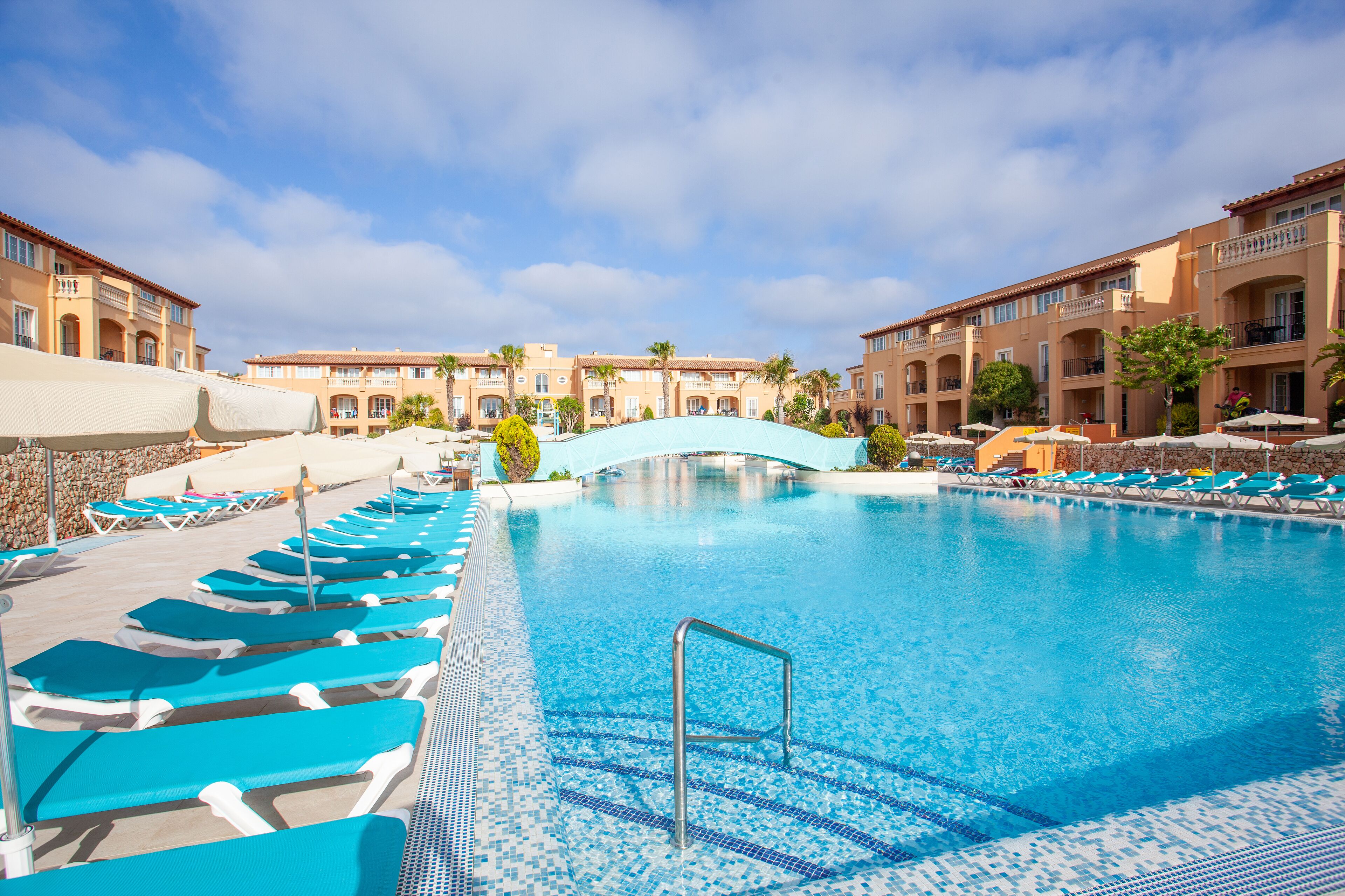 A swimming pool with blue lounge chairs and a bridge, surrounded by multi-story buildings with balconies.