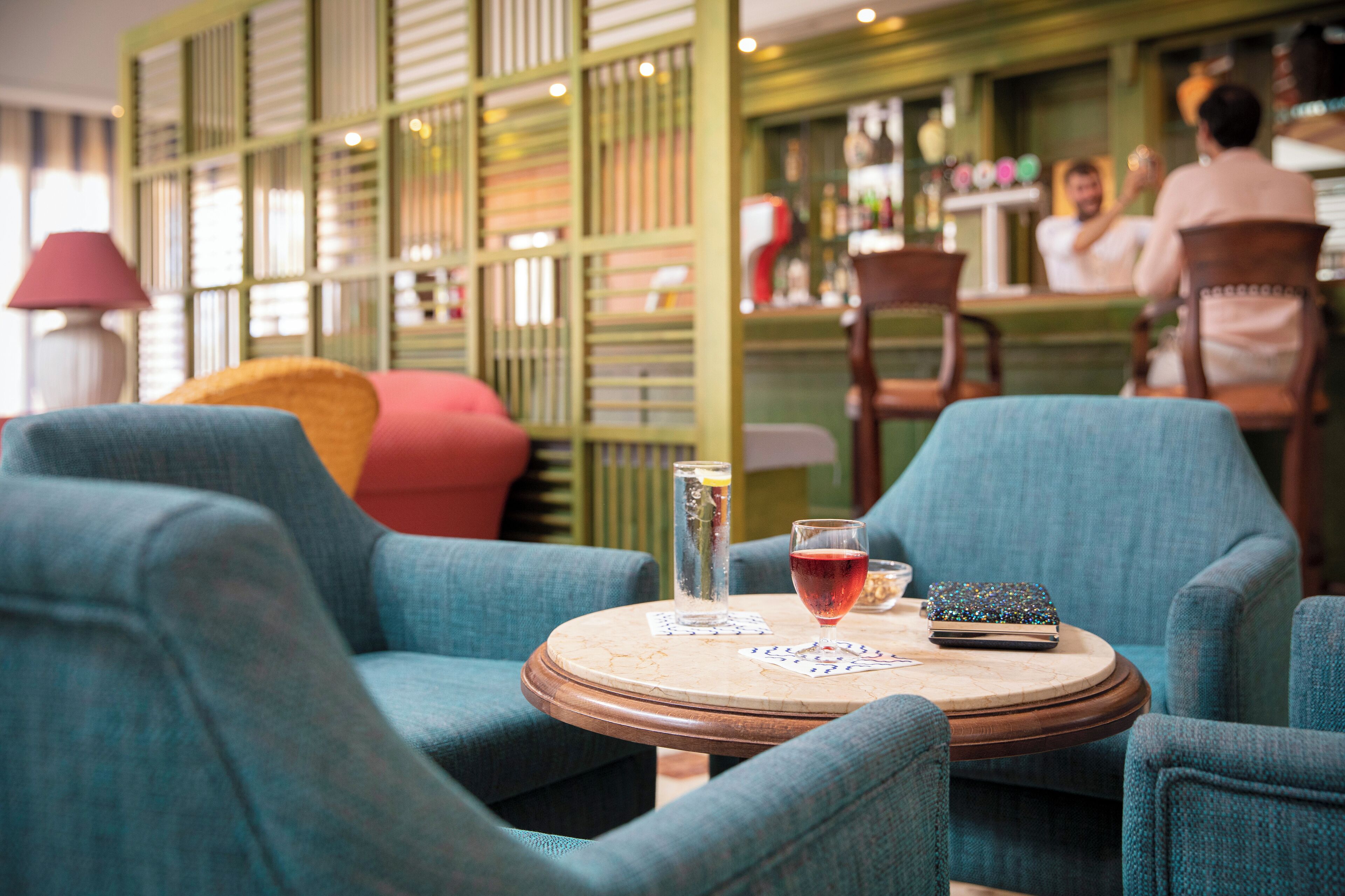 A round wooden table with a blue upholstered chair, a glass of red drink, playing cards, and a small blue bag.