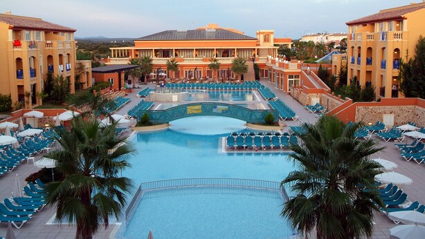 A large swimming pool with multiple lounge areas and palm trees, surrounded by hotel buildings with balconies and outdoor seating. - Grupotel Club Turquesa Mar (Ciutadella de Menorca)