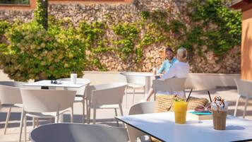 Um café ao ar livre com mesas e cadeiras, uma parede de pedra com vegetação e um céu claro.