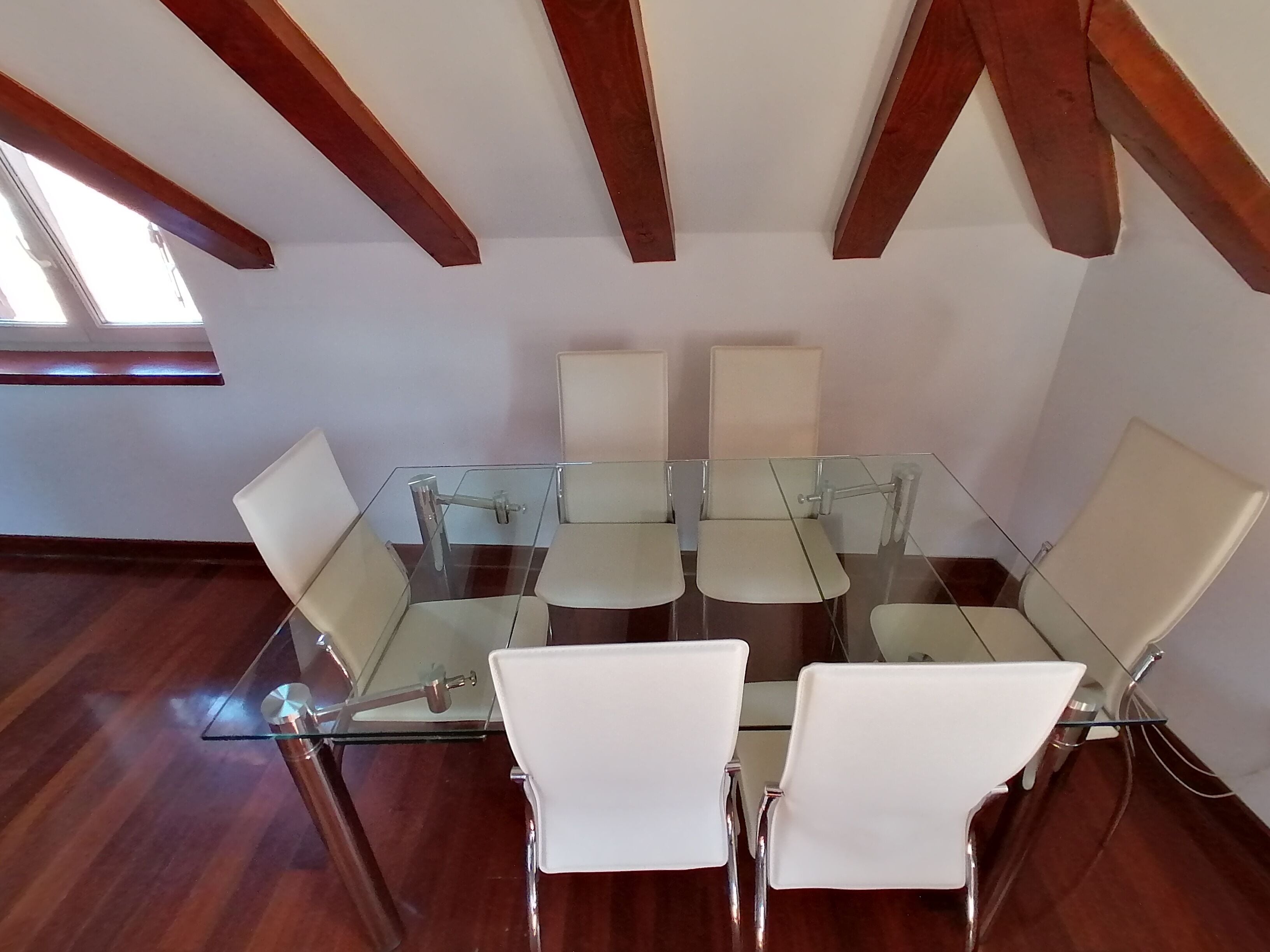 A conference room with a glass table and white chairs, wooden beams on the ceiling, and a window providing natural light.